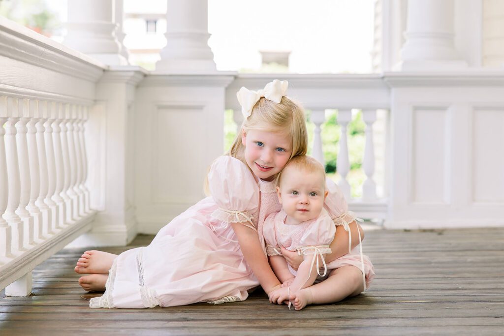 Two young sisters in matching pink heirloom dresses sitting together on a white porch during a family milestone session in Bainbridge, GA