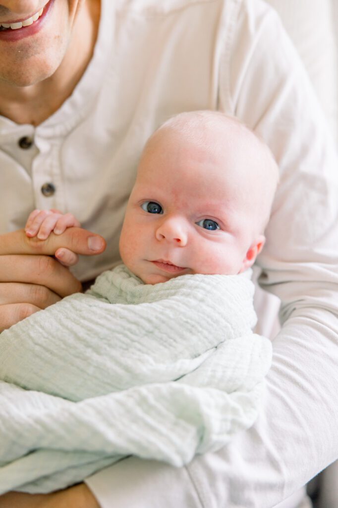 Tallahassee newborn photographer Sarah Gray captures a wide-eyed newborn boy wrapped in a soft swaddle, held by his father.